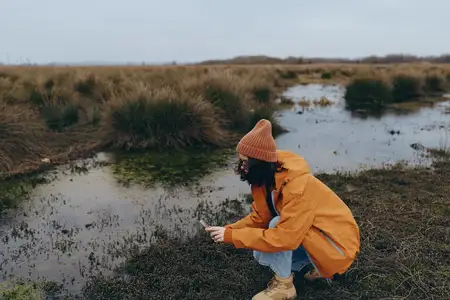 Женщина на природе в повседневном стиле: плащ и шляпа под облачным небом, стоит на коленях возле пруда на заболоченной территории