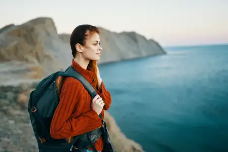 Путешественница в свитере и с рюкзаком за спиной на фоне гор и океана