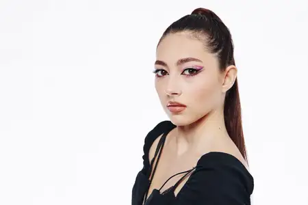 Портрет модной молодой женщины с аккуратно зачесанным назад хвостом, яркой розовой подводкой для глаз и глянцевыми губами, одетой в стильный черный топ на простом белом фоне