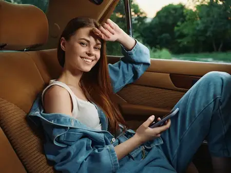 Счастливая молодая женщина, одетая в джинсовую одежду, держит смартфон, улыбаясь, сидя в автомобиле на фоне пышной зелени, идеально подходит для тем радости и спонтанности