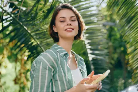 Женщина на улице среди зелени, нежная улыбка, полосатая рубашка, простые аксессуары, мягкий свет, естественный момент, теплая атмосфера, свежий воздух, уверенное и расслабленное настроение, портрет…