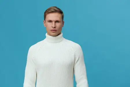 Уверенный молодой человек с нейтральным выражением лица, одетый в белую водолазку, позирует на фоне сплошного синего фона