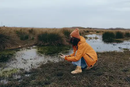 Женщина на отдыхе на природе: стоит на коленях в дождевике и шапке, кормит уток хлебом на илистом берегу
