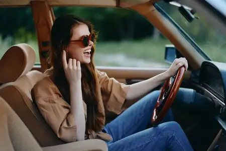 Веселая молодая женщина в солнцезащитных очках и повседневной одежде смеется за рулем винтажного автомобиля, передавая суть свободы и приключений на фоне пышной зелени, что идеально подходит для…