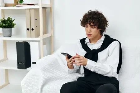 Кудрявый парень сидит на белом диване со смартфоном в руках