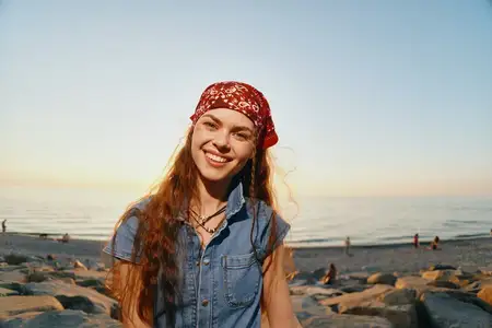 Женщина в стиле лайфстайл, одетая в современный бохо-вестерн деним в уличном стиле и красную бандану, радостно улыбается на каменистом океанском берегу в золотой час, запечатленная в теплых цветах…