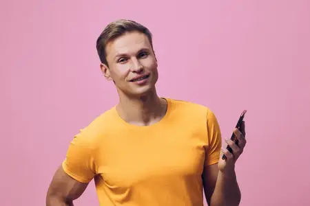 Улыбающийся мужчина со смартфоном в оранжевой футболке стоит на розовом фоне, держа мобильный телефон, что демонстрирует концепцию цифрового кочевника и технологии удаленной работы и общения