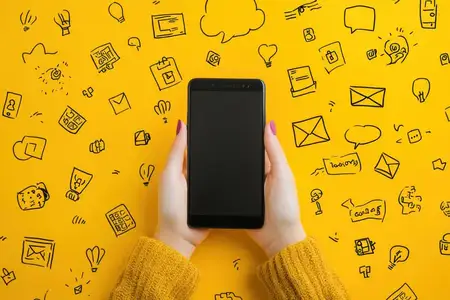 Крупный план рук, держащих смартфон на ярком желтом фоне, в окружении рисунков, символизирующих технологии и общение