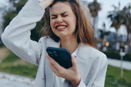 Молодая женщина испытывает стресс, держа смартфон с новостями о финансовом кризисе, осенний уличный фон, светло-серый блейзер, эмоциональное выражение лица