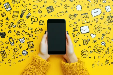 Крупный план рук, держащих смартфон на ярком желтом фоне, заполненном рисунками, изображающими темы общения, технологий и социальных сетей