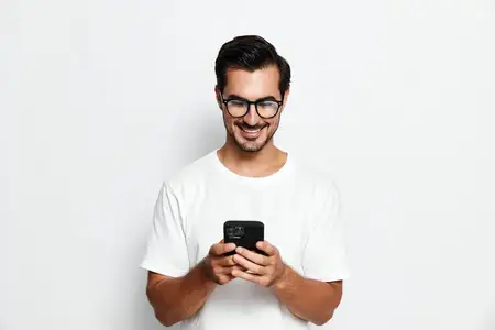 Весёлый молодой человек в очках улыбается, пользуясь смартфоном, демонстрируя современные технологии