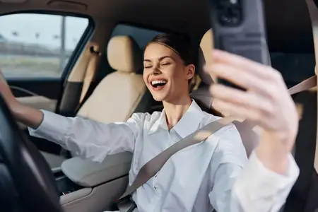 Женщина на водительском сиденье автомобиля делает обычное селфи на мобильный телефон, запечатлевая момент повседневной жизни и современных технологий