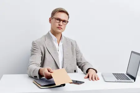 Серьезный мужчина работает с ноутбуком и смартфоном, держит в руке визитную карточку, одет в серую куртку, белую рубашку и очки, сидит за белым столом, изолирован на белом фоне