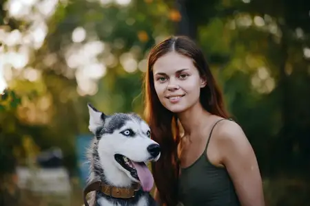 Женщина с собакой породы хаски улыбается и ласково гладит своего любимого пса во время прогулки на природе в парке осенью на фоне заката