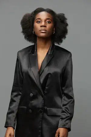 На этом изображении представлена уверенная африканская женщина в черном сатиновом блейзере, подчеркивающем ее натуральные кудри и самообладание на нейтральном сером фоне