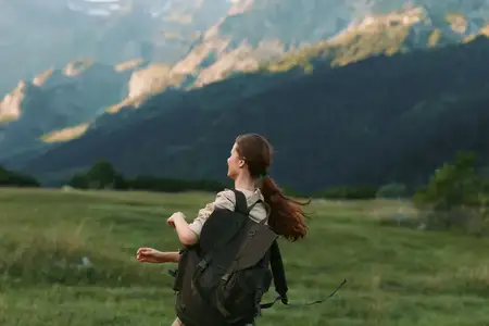 Спокойная, активная outdoor-сцена с женщиной-туристкой в одиночестве с рюкзаком на травянистом лугу возле высоких гор, подчеркивающая природу, исследование, приключения и хорошее самочувствие при…