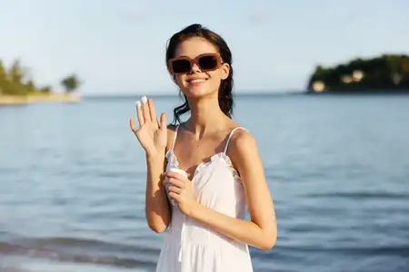 Улыбающаяся женщина в белом платье машет рукой, стоя на солнечном пляже, в солнцезащитных очках, наслаждаясь теплой летней погодой на фоне спокойной воды