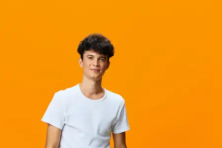 Молодой человек с кудрявыми волосами и теплой улыбкой в белой футболке уверенно позирует на ярко-оранжевом фоне, излучая позитив и расслабленную летнюю атмосферу
