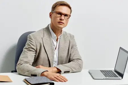 Сосредоточенный мужчина средних лет в очках, бежевом пиджаке и белой рубашке сидит за офисным столом с ноутбуком и смотрит прямо в камеру с серьезным выражением лица, профессиональная обстановка