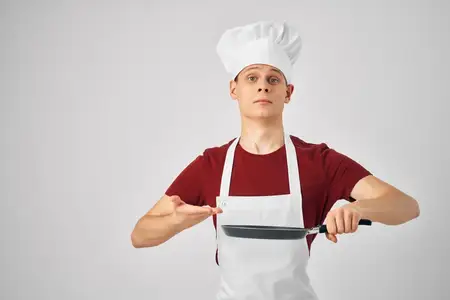 Повар-мужчина в белом фартуке держит в руке сковороду и готовит еду