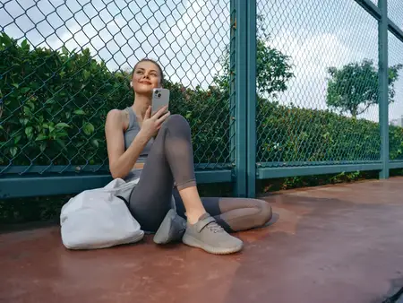 Подтянутая молодая женщина в спортивной одежде отдыхает, сидя на поверхности теннисного корта и пользуясь смартфоном