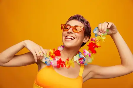 Портрет женщины с цветочным гавайским венком на шее, которая весело танцует и улыбается в яркой одежде на оранжевом фоне, в солнцезащитных очках