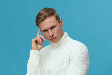 Портрет сосредоточенного молодого человека с blond-волосами, в белом свитере с высоким воротником, пристально смотрящего в камеру с рукой у виска, на однородном голубом фоне, студийная обстановка