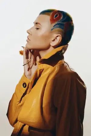 Профиль модной женщины, демонстрирующий яркую выбритую прическу и креативные цвета, одетой в текстурированную желтую кожаную куртку, на чистом студийном фоне, подчеркивающий современный стиль и…