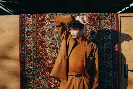 Модный молодой человек уверенно стоит на фоне богатого, красочного ковра, подчеркивая современный стиль и элегантность