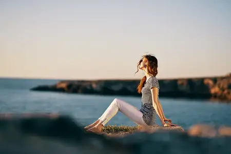 Женщина в полосатой футболке и белых брюках сидит на пляже у моря в горах