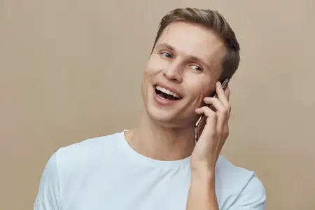Улыбающийся молодой светлокожий мужчина разговаривает по телефону, держит мобильное устройство, одет в белую футболку на бежевом фоне, счастливое выражение лица