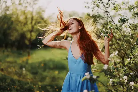 Женщина с красивой улыбкой, обнажающей зубы, и длинными развевающимися на ветру волосами танцует на закате в парке рядом с цветущими деревьями