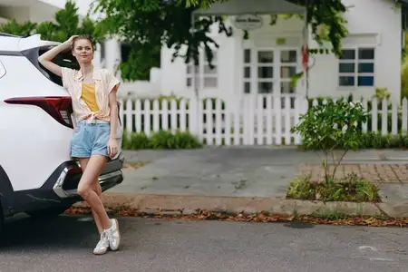 Молодая женщина в повседневной одежде стоит рядом с белым автомобилем, излучая уверенность и радость