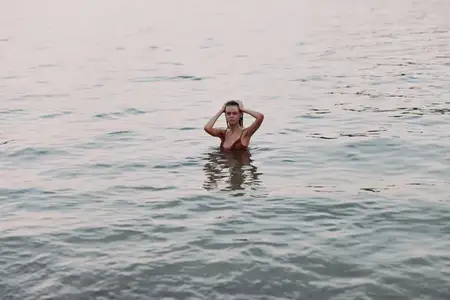Женщина в купальнике стоит одна в спокойной воде на закате, запустив руки в волосы, передавая безмятежность и созерцание в безмятежной пляжной обстановке