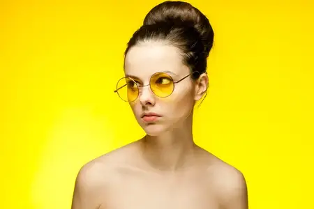 Женщина с обнаженными плечами, в желтых очках, держит руки возле лица