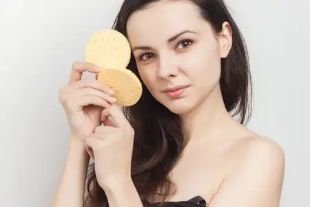 Привлекательная брюнетка с обнаженными плечами держит спонжи возле лица, демонстрируя чистую кожу