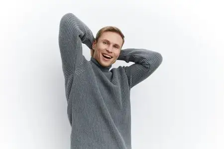 Весёлый молодой человек со светлыми волосами, одетый в тёплый серый вязаный свитер, радостно улыбается в камеру, выражая счастье и уверенность