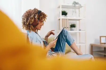 Счастливая женщина дома: отдыхает под музыку и играет на планшете, сидя на уютном диване в гостиной
