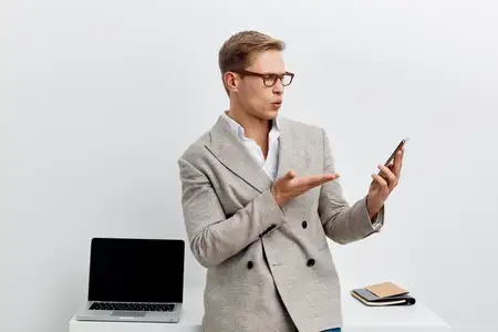Удивленный мужчина пользуется смартфоном рядом с ноутбуком на белом столе