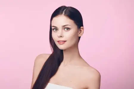 Привлекательная молодая женщина с длинными каштановыми волосами позирует на розовом фоне, накинув на голову белое полотенце