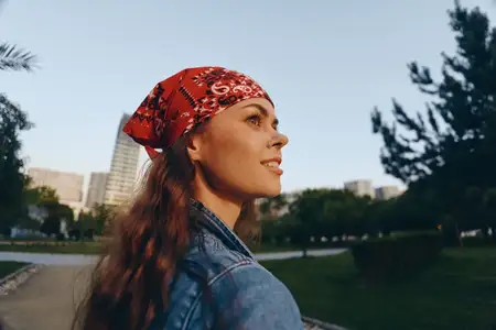 Молодая женщина в красной бандане и джинсовой куртке задумчиво смотрит вдаль, стоя в солнечном городском парке в окружении зеленых деревьев и небоскребов