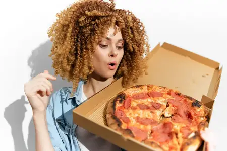 Радостная молодая женщина с вьющимися волосами смотрит на вкусную пиццу в коробке на вынос, демонстрируя восторг