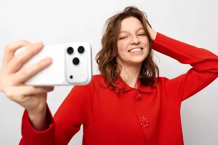 Счастливая девушка в красном пуловере делает селфи на мобильный телефон, касаясь волос и улыбаясь на белом фоне