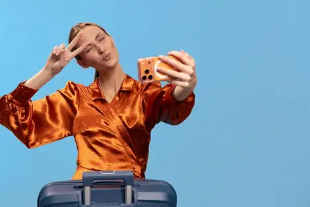 Улыбающаяся женщина в яркой оранжевой атласной блузке держит смартфон для селфи, показывая жест «мир» над синим чемоданом