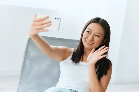 Наслаждающаяся, улыбающаяся, загорелая, милая молодая азиатка машет рукой, приветствуя друга в прямом эфире во время видеозвонка-селфи дома