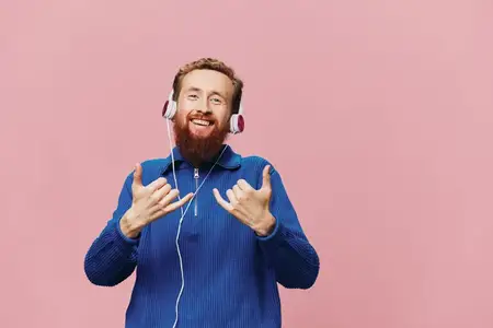 Портрет рыжеволосого мужчины в наушниках, который улыбается и танцует, слушая музыку на розовом фоне