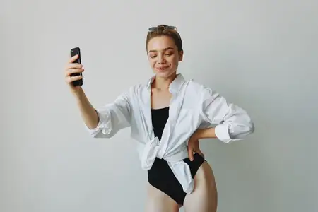 Подросток-девушка-блогер фотографирует и снимает себя на видео на свой телефон для контента в социальных сетях, видеоблогер, селфи на фоне белой стены с улыбкой дома, фрилансер