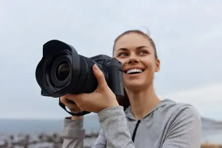 Радостная молодая женщина с фотоаппаратом, ярко улыбаясь, стоит на облачном пляже, готовая запечатлеть потрясающие моменты прибрежной фотографии