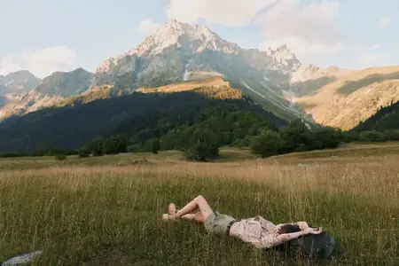 На ней лёгкое цветочное платье, она отдыхает среди полевых цветов, травы колышутся, а горы возвышаются позади; спокойная outdoor-сцена, передающая свободу, здоровье и естественный образ жизни