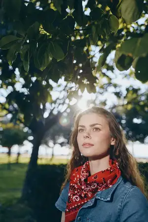 Молодая женщина в красном шарфе и джинсовой куртке мирно стоит под ветвями деревьев, сквозь которые пробиваются солнечные лучи, среди ярко-зелёных листьев, снятая в естественной парковой обстановке…